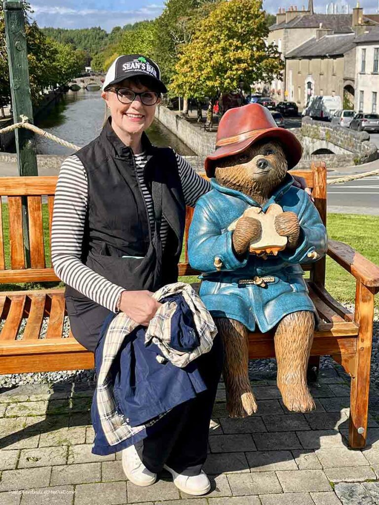 A woman sitting on a bench beside a statue of Paddington Bear eating a sandwich